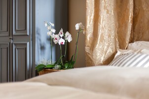 Una cama con ropa blanca, una cesta de mimbre con plantas verdes, y una pared con una puerta y cortinas.