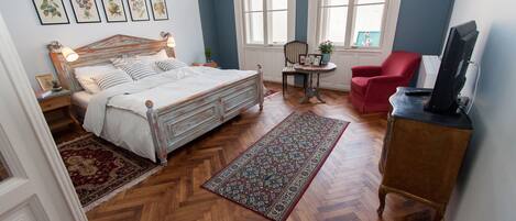 Un dormitorio con una cama de madera, una silla roja, una mesita con una planta y un televisor sobre un mueble de madera.