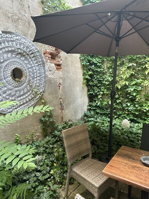 A wicker chair, a table, and a large umbrella in a garden area with a wall and greenery. - Camino Home (Cluj-Napoca)