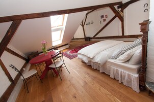 Un dormitorio con techo inclinado, vigas de madera, una cama con ropa blanca, una mesa roja con sillas y un jarrón con flores.