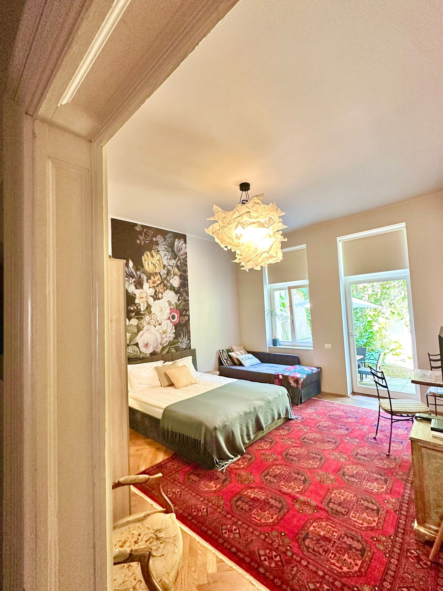 A bedroom with a large bed, a hanging light fixture, a floral wall mural, and a view of the outdoors through a window.