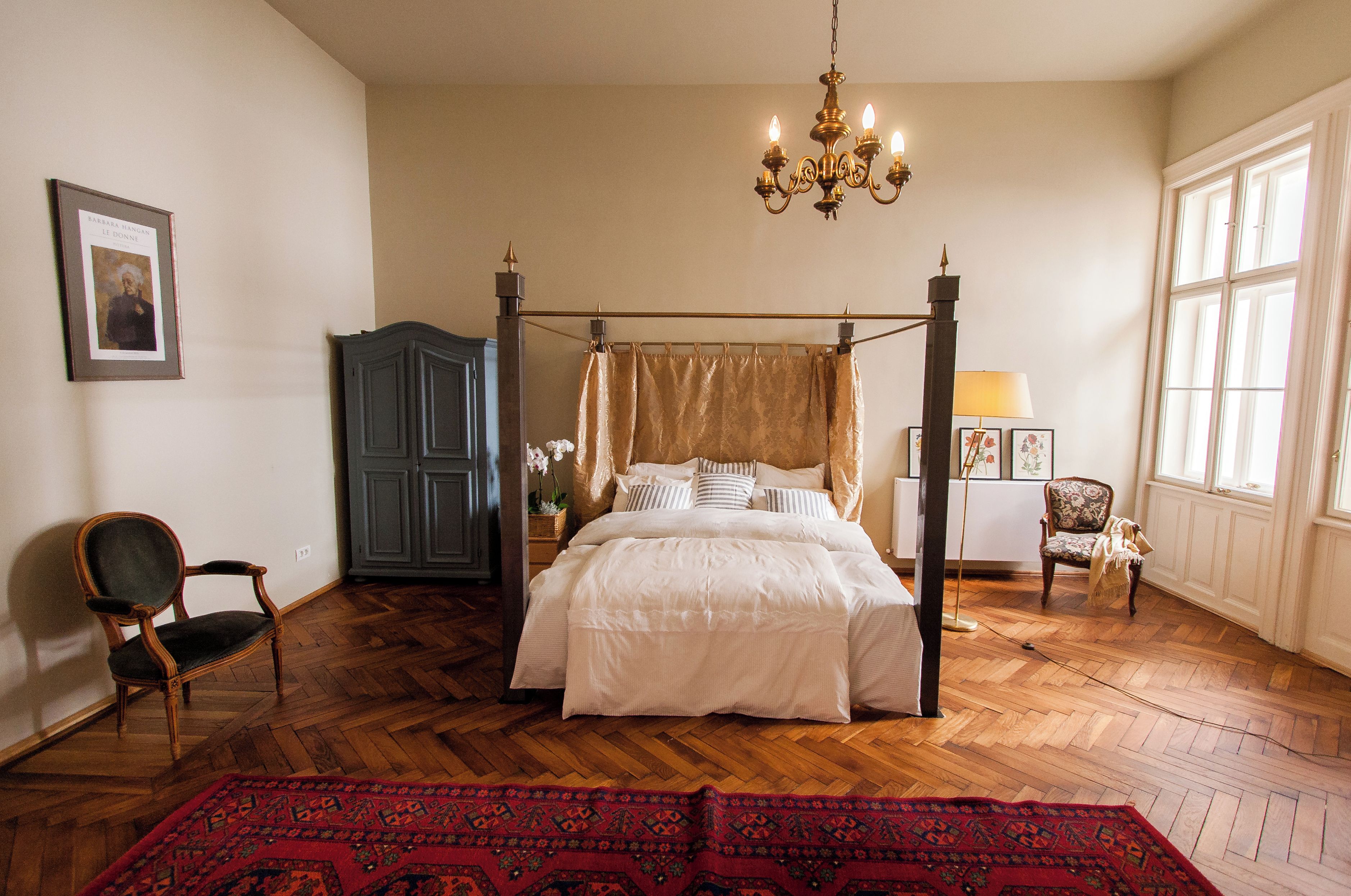 A four-poster bed with a canopy, a chandelier, a wooden floor, and a red patterned rug.