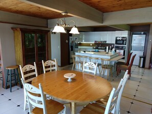 A dining area with a wooden table and white chairs, a kitchen with modern appliances, and a large window with curtains.