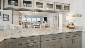 A spacious kitchen with marble countertops, grey cabinetry, and a dining area with white chairs. - Beach Condo Miles-long Views Sand and Sea, Access to Private Fishing Pier by RedAwning (Fernandina Beach)