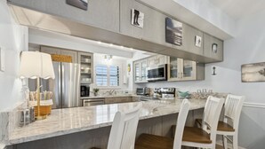 A modern kitchen with a large island, white bar stools, stainless steel appliances, and a marble countertop. - Beach Condo Miles-long Views Sand and Sea, Access to Private Fishing Pier by RedAwning (Fernandina Beach)