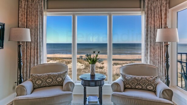 A room with two armchairs facing a large window overlooking a beach. A small table with a vase of flowers is between the chairs.