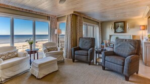 A spacious living room with a large window overlooking the beach, a ceiling fan, and a cozy seating area with a chair and ottoman.