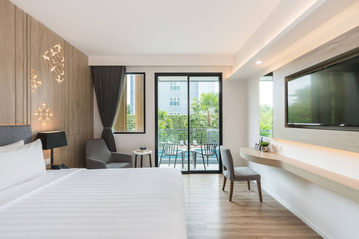 Ein modernes Hotelzimmer mit einem großen Bett, einer Sitzecke mit Tisch, einem an der Wand montierten Fernseher und einem Balkon mit Blick auf den Pool.