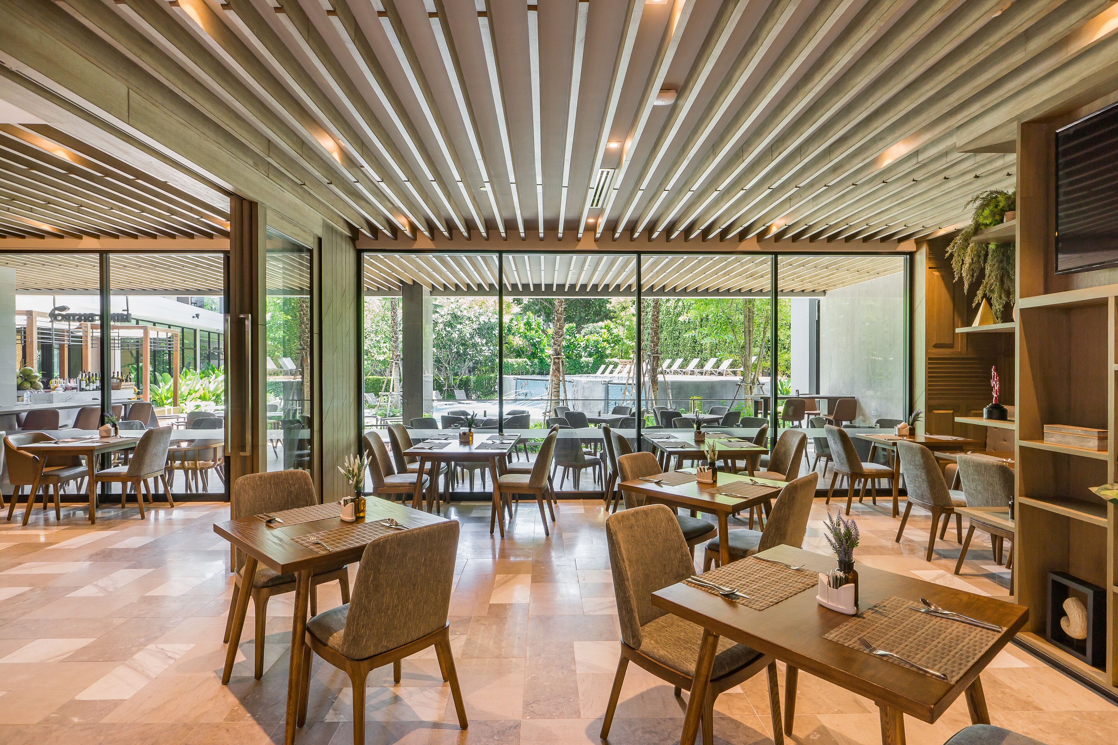 A modern restaurant with wooden tables and chairs, large windows, and a ceiling with wooden slats.