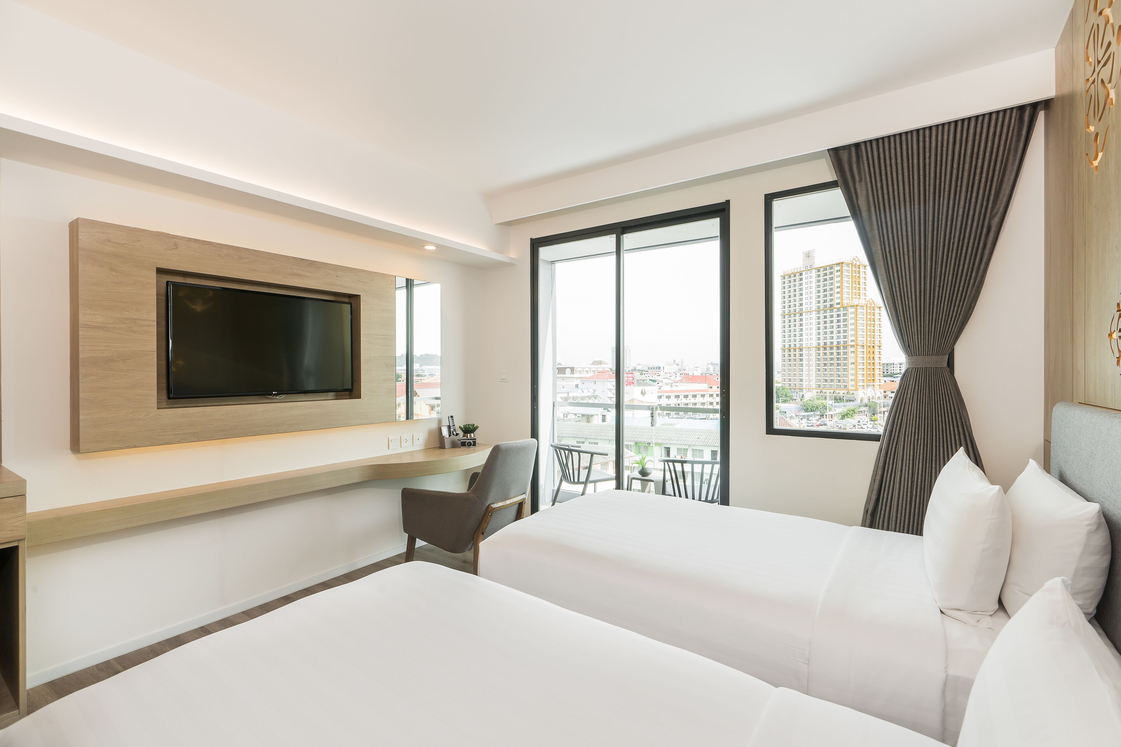 Een hotelkamer met twee bedden, een aan de muur bevestigde televisie, een bureau en uitzicht op de stad door grote ramen.