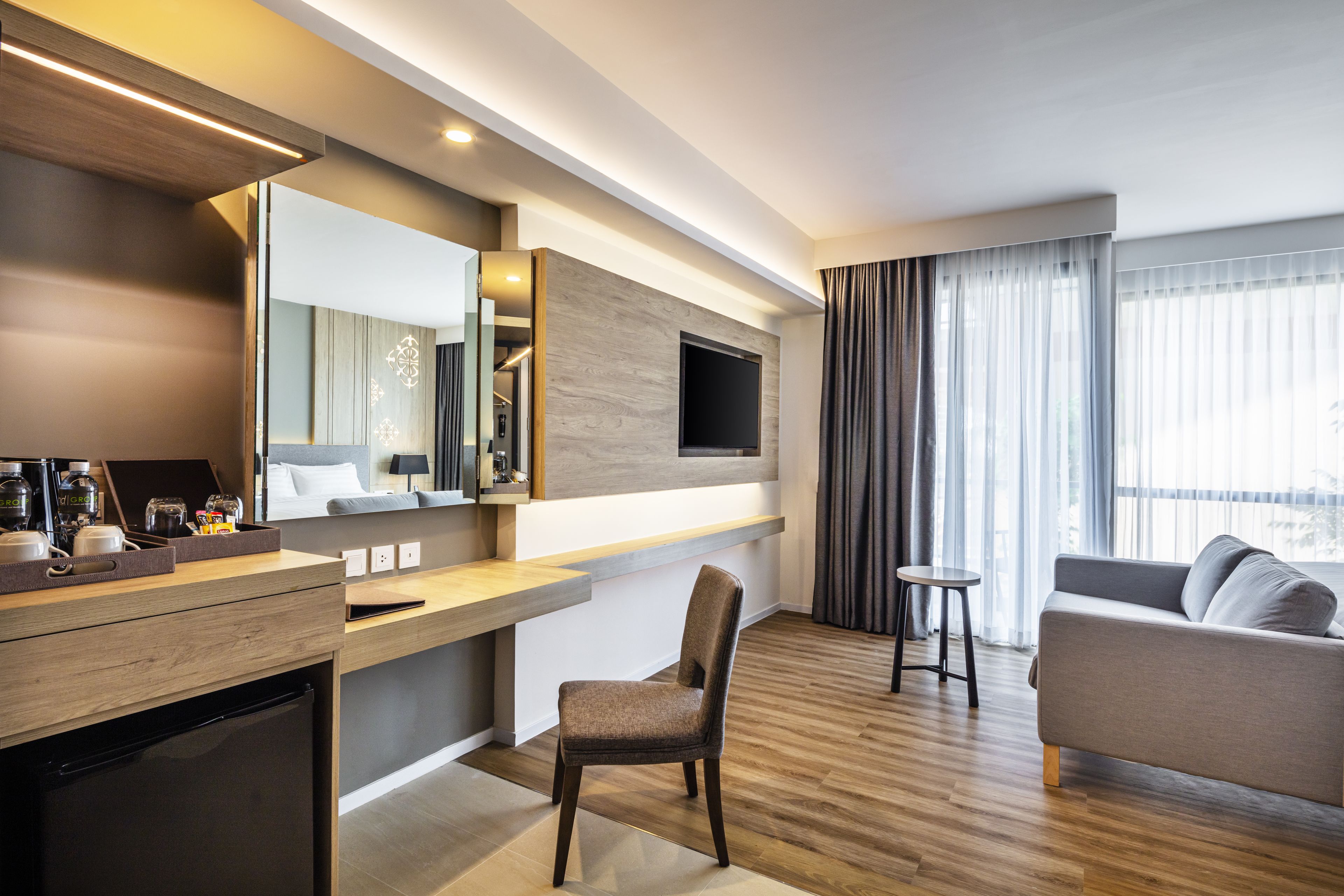 A modern hotel room with a wooden floor, a sofa, a dining area with a table and chairs, a kitchen area with a refrigerator, and a large mirror.