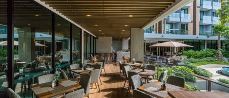 Un espace de restauration extérieur moderne, doté de tables et de chaises en bois, de grandes fenêtres et offrant une vue sur l’intérieur du bâtiment.