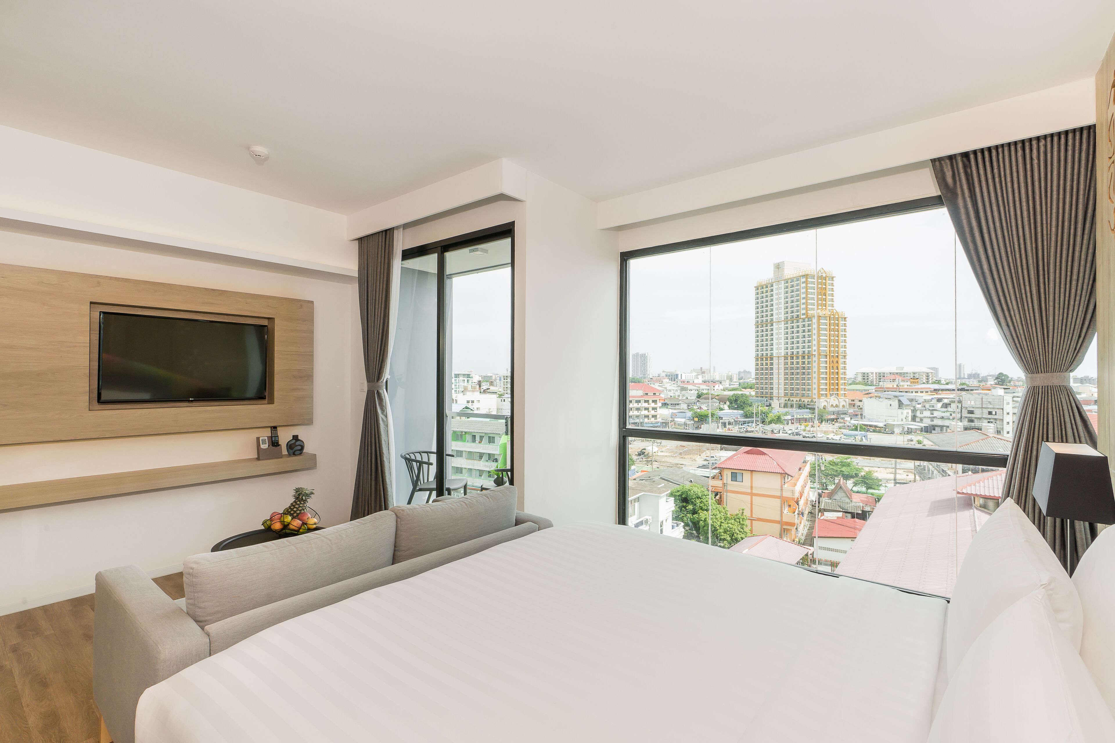 A hotel room with a bed, a TV mounted on the wall, a window with curtains, and a view of the cityscape.