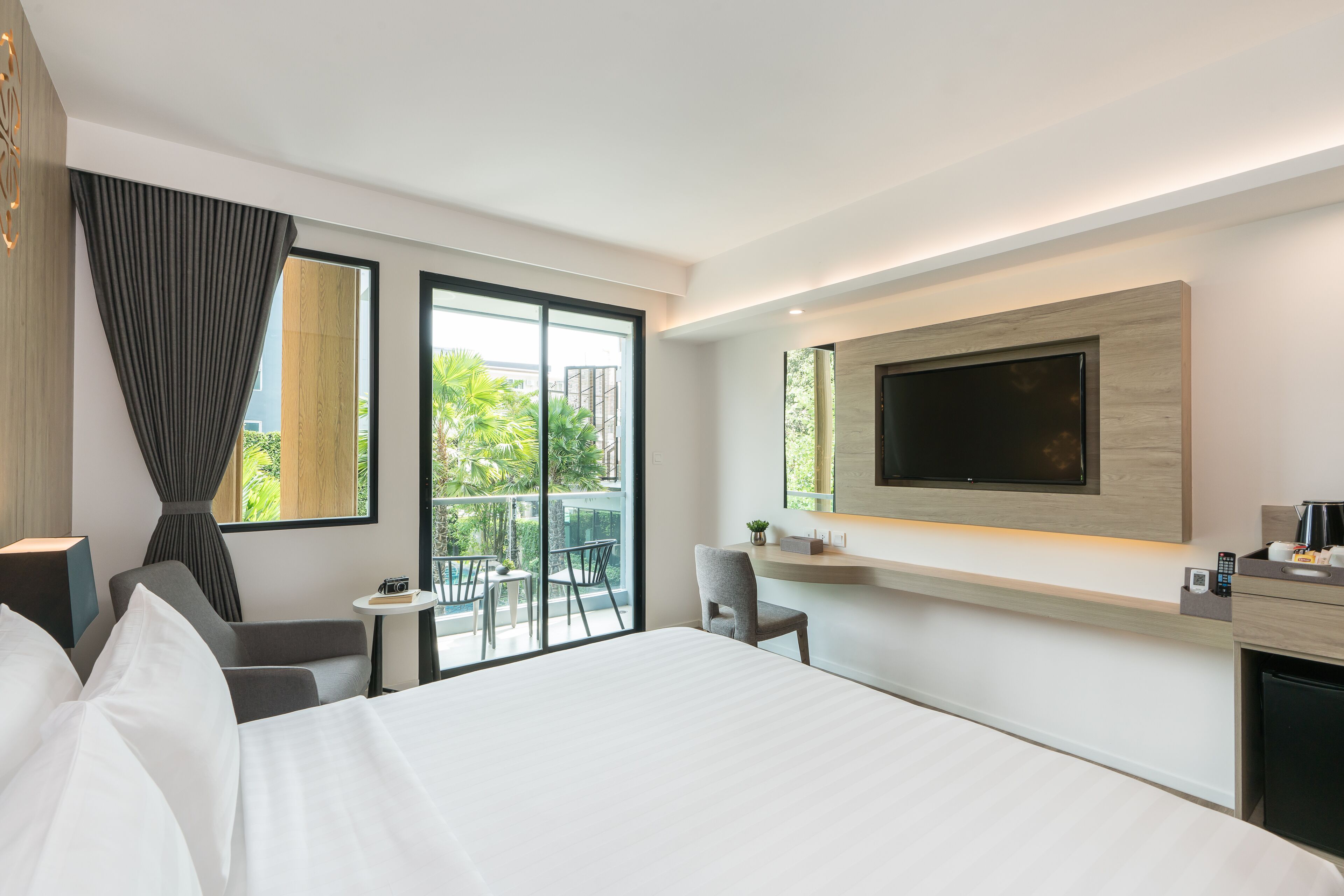 A modern hotel room with a large bed, a flat-screen TV mounted on the wall, a small table with a chair, and a balcony with outdoor seating.
