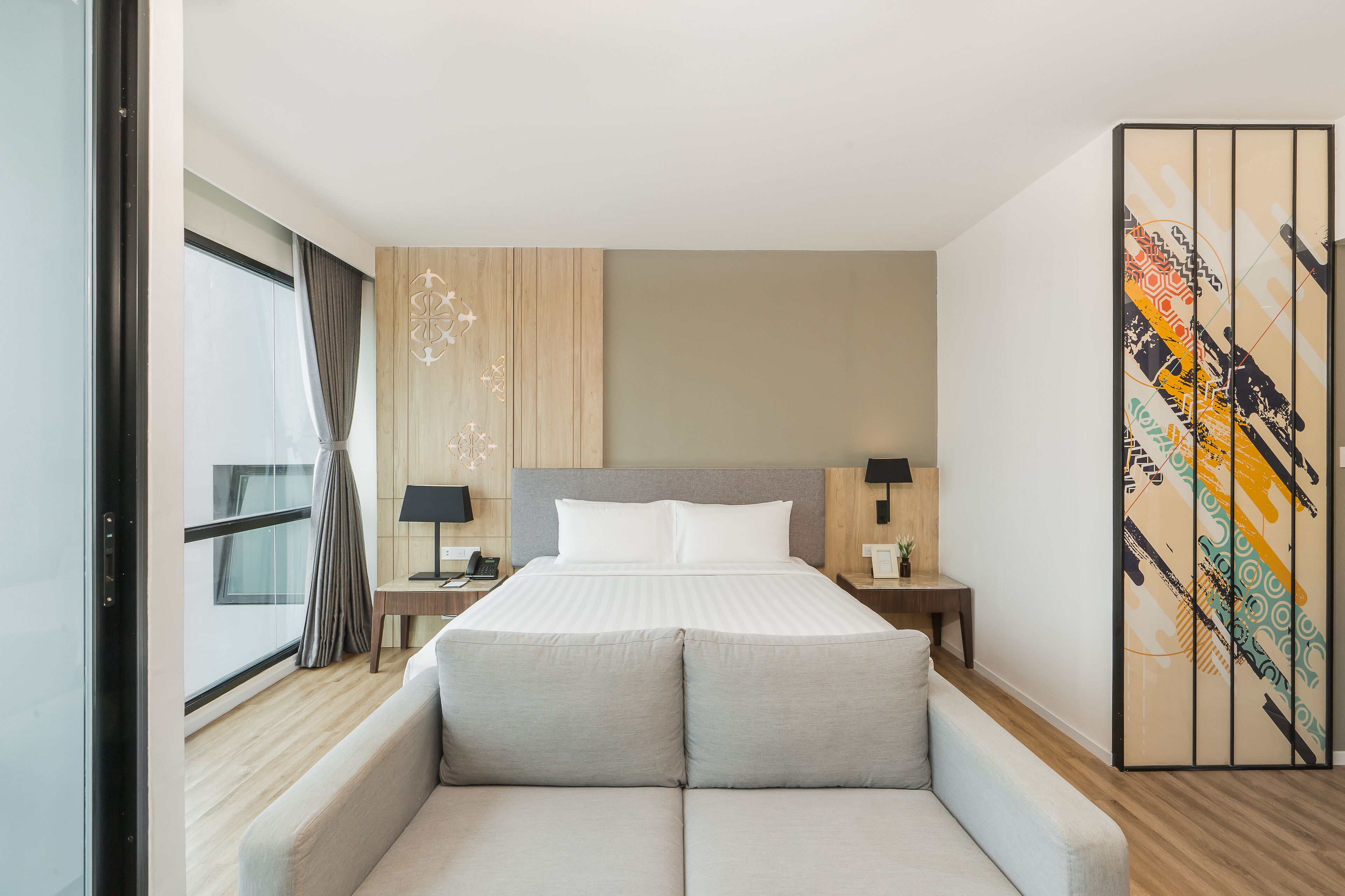 A modern hotel room with a large bed, a sofa, a decorative wall panel, and a window with curtains.