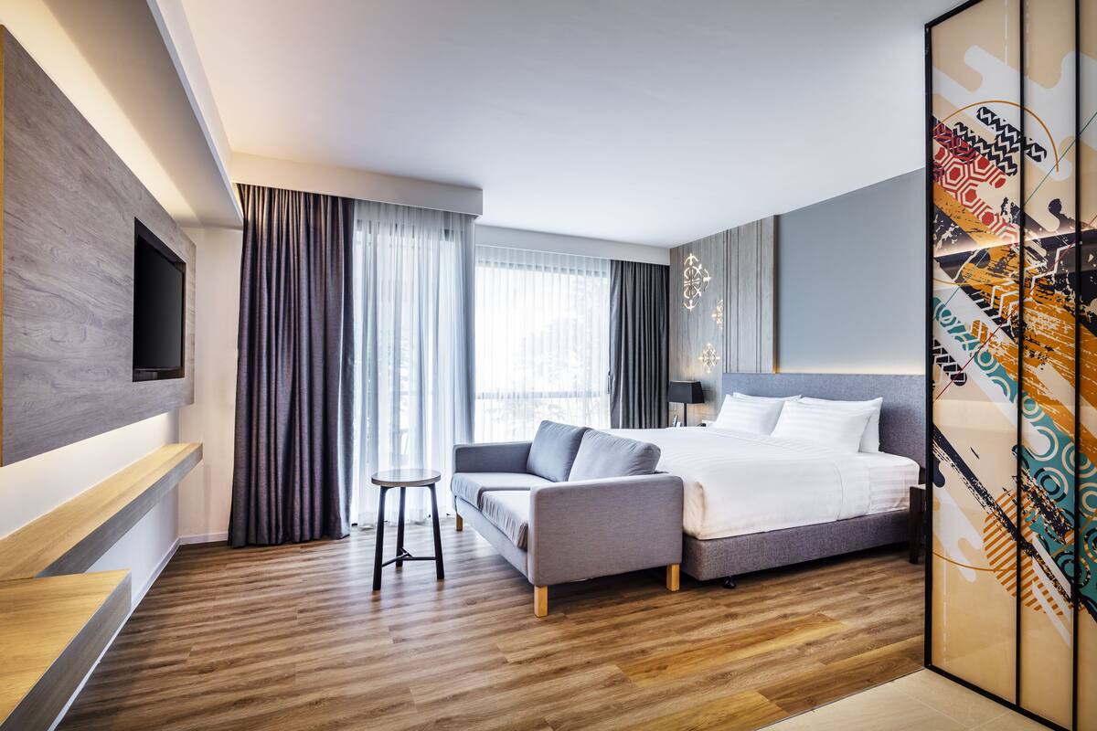 Ein modernes Hotelzimmer mit einem großen Bett, einer Sitzecke, einem an der Wand montierten Fernseher und einem großen Fenster mit Vorhängen.