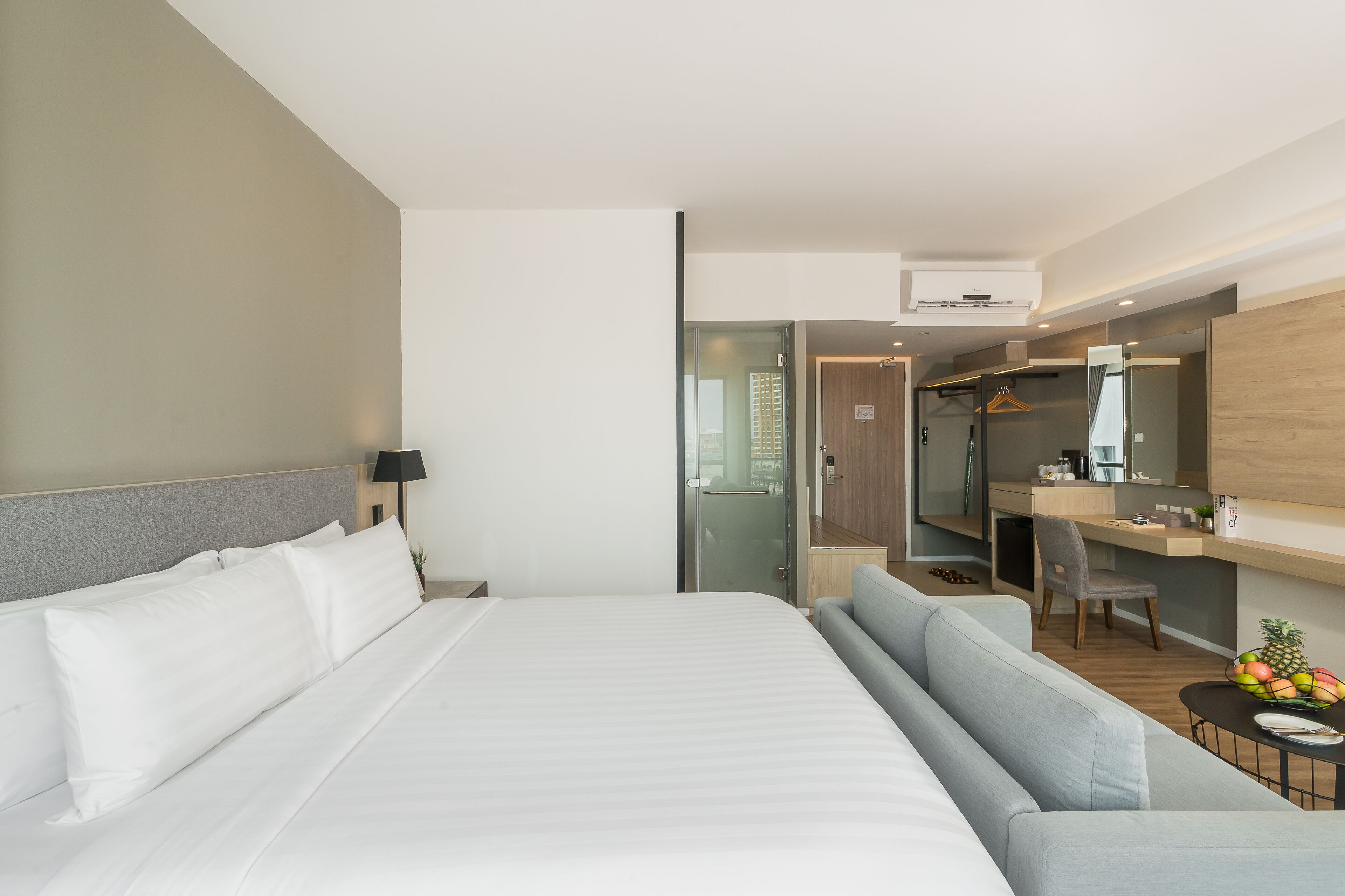 A modern hotel room with a large bed, a desk, a chair, a small table with fruit, and a mirror.