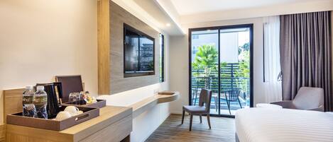 A hotel room with a wooden desk, a bed, a TV, and a balcony with a view of greenery.