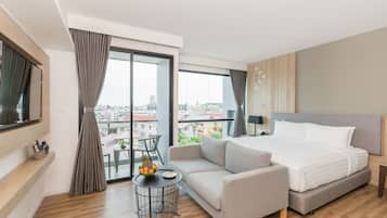 Quarto de hotel moderno com uma cama grande, um sofá, uma mesinha redonda com frutas e vista para a cidade através de amplas janelas.