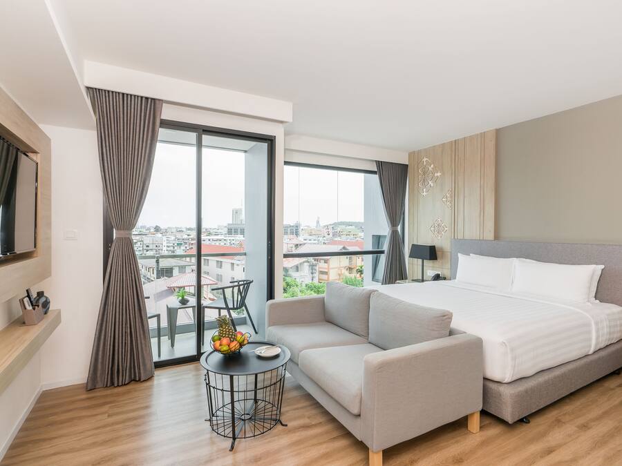 Quarto de hotel moderno com uma cama grande, um sofá, uma mesinha redonda com frutas e vista para a cidade através de amplas janelas.