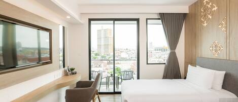 A modern hotel room with a large bed, a TV mounted on the wall, a balcony with a view of the city, and a seating area with a chair and table.