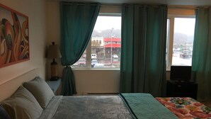 A hotel room with a bed, a TV, a window with curtains, and a painting on the wall. - Aquamarine Upbeat Studio on the Mall  (Kodiak)