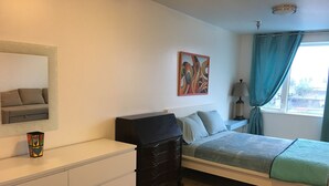 A bedroom with a bed, a dresser, a nightstand, a lamp, a painting, and a window with curtains. - Aquamarine Upbeat Studio on the Mall  (Kodiak)