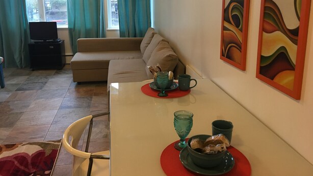 A living room with a sofa, a dining table with a red placemat, a TV, and abstract paintings on the wall. - Aquamarine Upbeat Studio on the Mall  (Kodiak)
