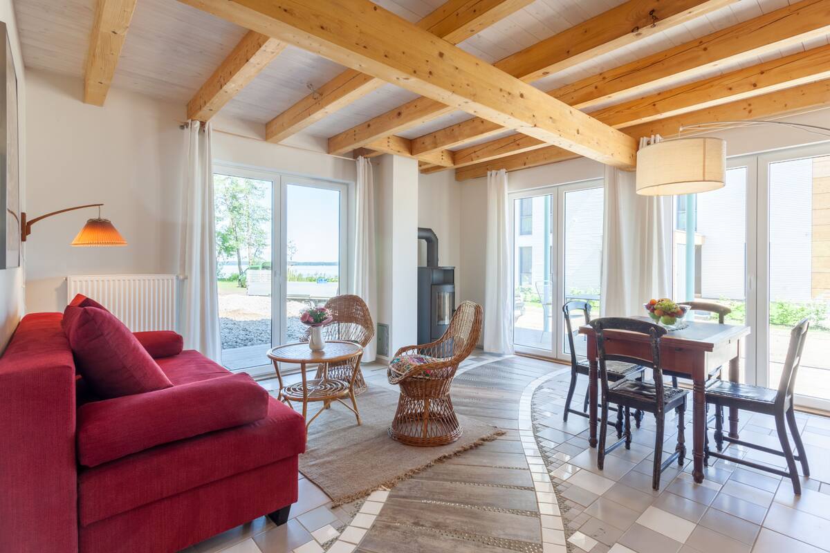 Uma sala de estar com um sofá vermelho, cadeiras de vime, uma mesa de jantar com cadeiras, uma lareira e janelas amplas.