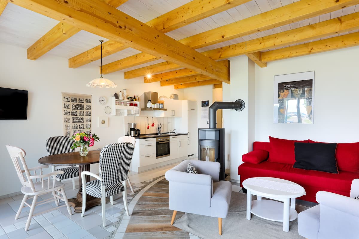 Uma sala de estar moderna com um sofá vermelho, poltronas brancas e uma área de jantar com mesa de madeira e cadeiras xadrez. Uma cozinha com armários brancos e um fogão é visível ao fundo.