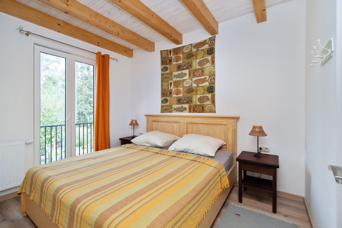 Quarto com cama, vigas de madeira, janela com cortinas, tapeçaria na parede e mesa de cabeceira com abajur.