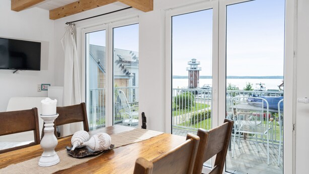 A dining area with a wooden table, chairs, and a candle. There is a TV mounted on the wall and a view of a balcony with a railing and a building in the background. - Plau Lagoons (Plau am See)