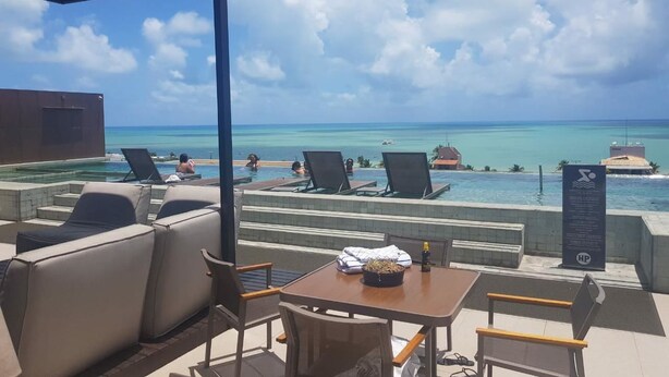 A poolside area with lounge chairs and a table, overlooking a clear blue sea and a resort building. - Holandas Prime (João Pessoa)