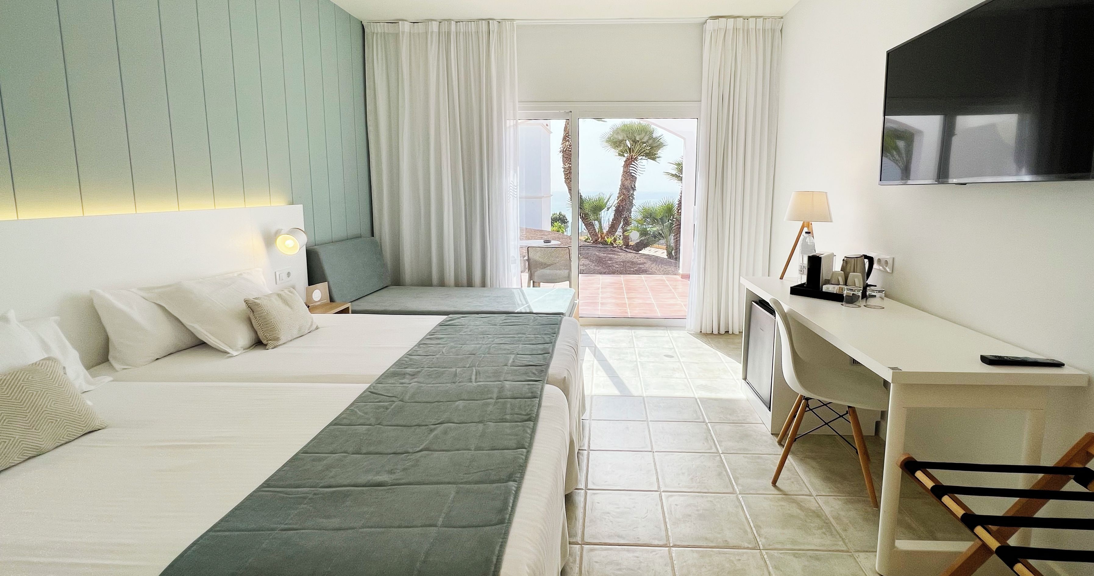 Habitació d'hotel amb dos llits, un escriptori, una televisió i vista a palmeres.