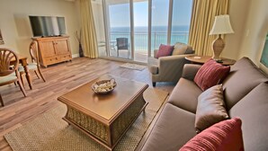 A spacious living room with a large window offering a view of the ocean, a ceiling fan, a television, and a cozy seating area with a sofa and armchairs.