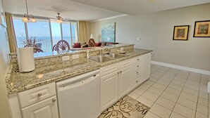 A modern kitchen with granite countertops, white cabinetry, and a dishwasher. There is a large window providing a view of the ocean.