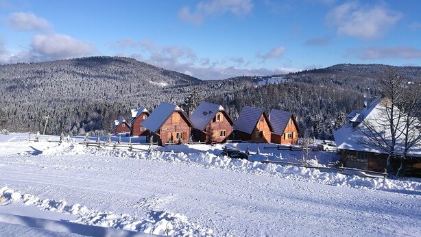 A row of wooden cabins with snow-covered roofs, set against a backdrop of snow-covered trees and mountains under a clear blue sky. - Lodge Nagramak (Bajina Bašta)