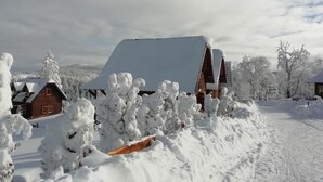 A snow-covered landscape with a wooden cabin and a large building with a sloped roof. - Lodge Nagramak (Bajina Bašta)