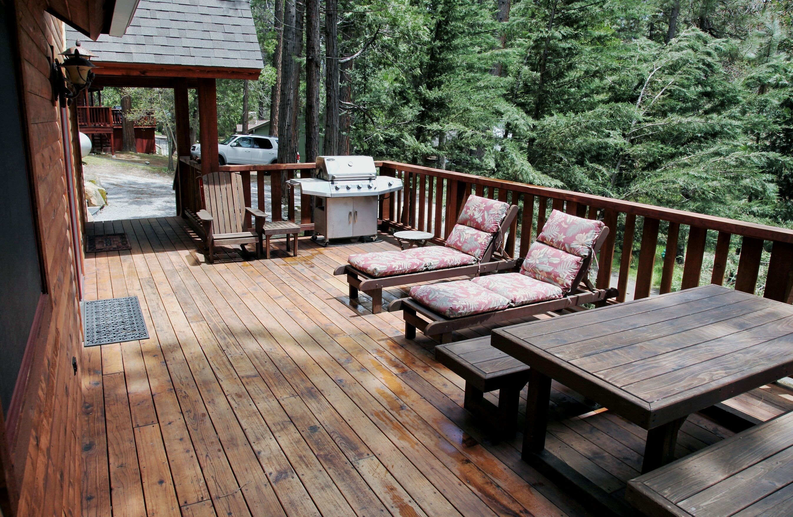 Una terraza de madera con mobiliario exterior, una parrilla y vista a un bosque.