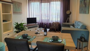A dining area with a wooden table set for two, a television, and a window with curtains. - Vacation apartment L' Ancora (mill view) (sint-annaland)