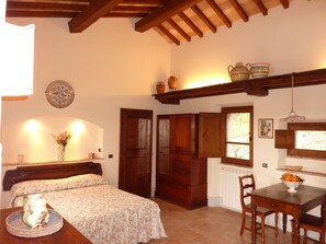 Une chambre à coucher avec un lit, une table de chevet et une petite table sur laquelle se trouve un saladier rempli de fruits. Une fenêtre offre une vue sur la verdure à l’extérieur.