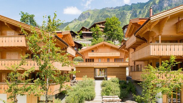 Una cabaña de estilo chalet, con balcones, rodeada de follaje y un paisaje montañoso.