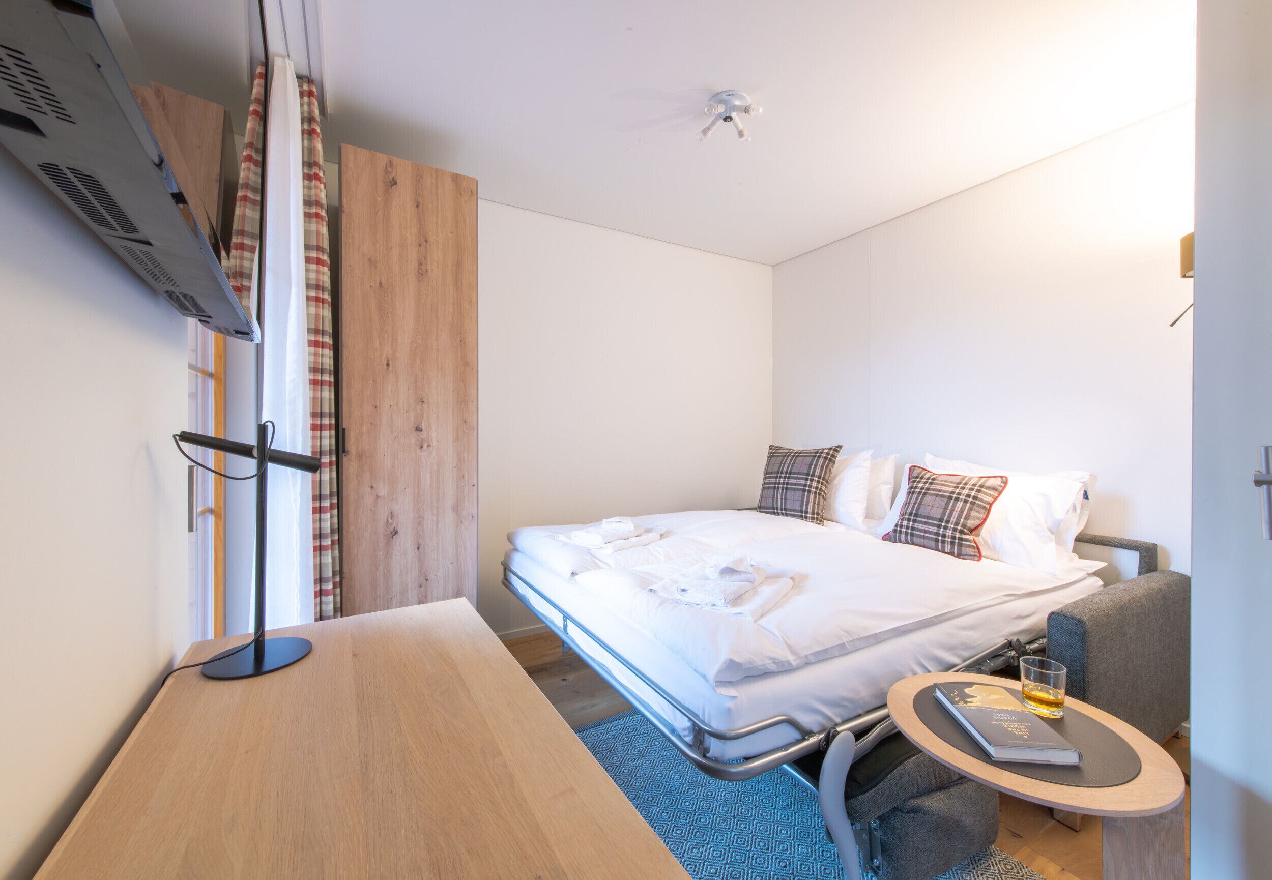 A compact hotel room with a bed, a small table with a drink, and a TV mounted on the wall.