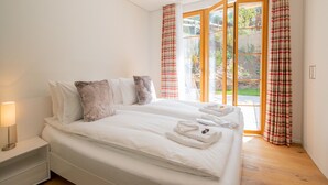 Una cama bien hecha con sábanas blancas y un almohadón mullido, una lámpara de noche y vistas a un jardín a través de ventanas amplias con cortinas a cuadros.
