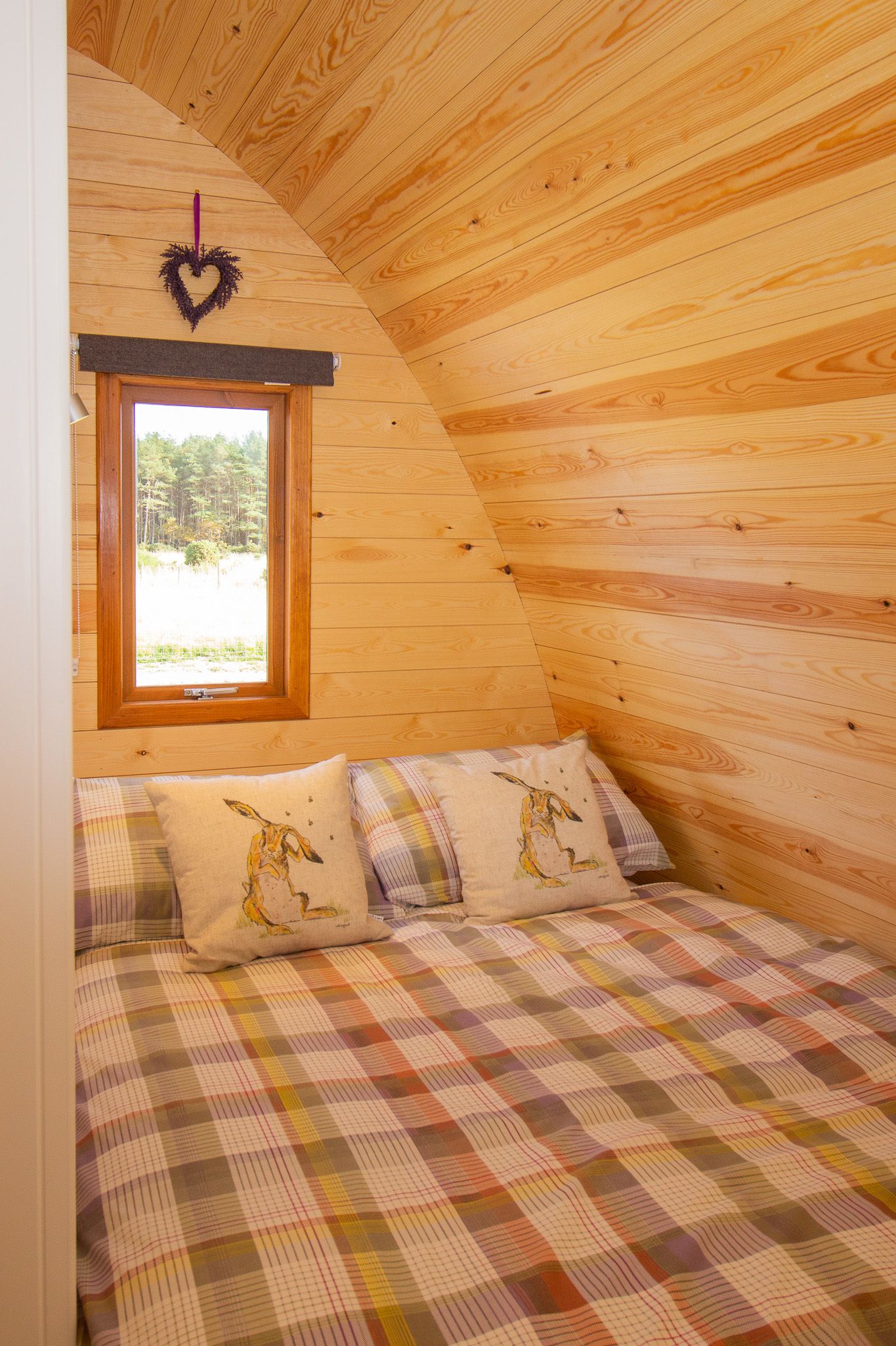 Ein gemütliches Blockhauskabinenzimmer mit kariertem Bett, einer herzförmigen Dekoration und einem Fenster mit Blick auf einen Wald.