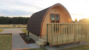 Una estructura de madera tipo cabaña con techo a dos aguas, una terraza y una puerta, rodeada por un camino de grava y césped.