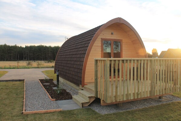 A wooden A-frame structure with a deck and a door, surrounded by a gravel path and grass. - Evelix Pods (Dornoch)