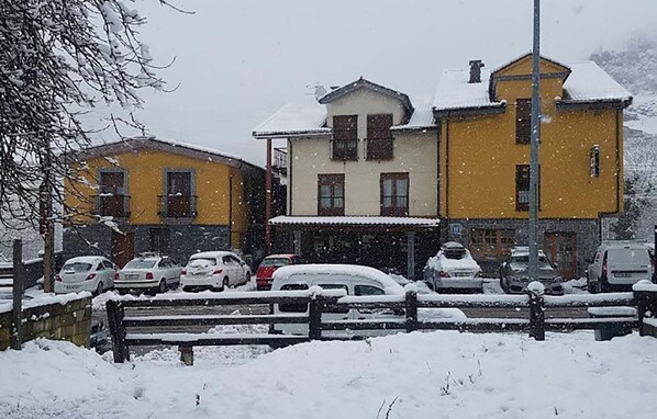 A two-story building with a yellow facade and a white roof, surrounded by parked cars and a snow-covered ground. - Hotel Peña Pandos (Aller)