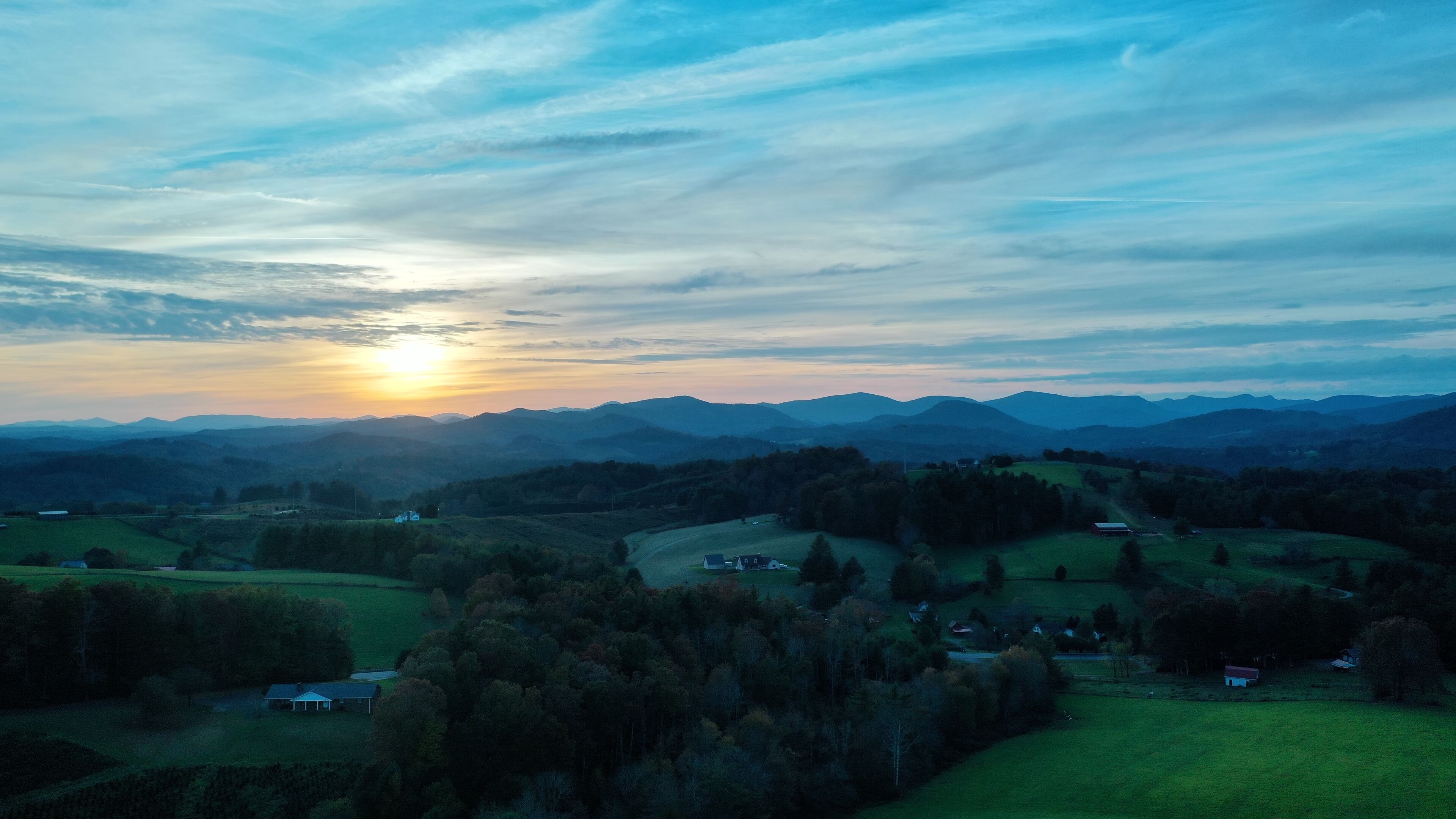 Une vue panoramique d’un paysage rural, avec des collines ondoyantes, des maisons dispersées et un coucher de soleil en arrière-plan.