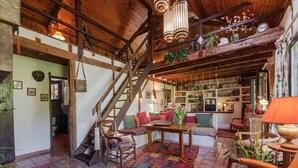 A rustic interior with exposed wooden beams, a staircase, a cozy seating area, and a well-stocked bookshelf.
